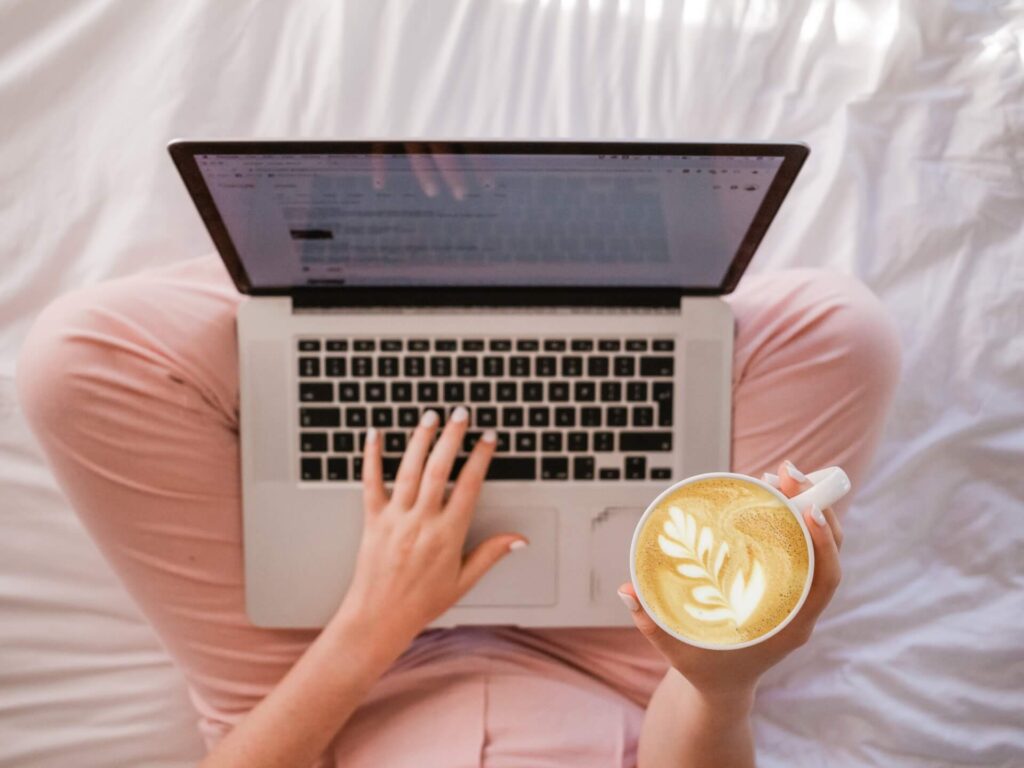 Woman sitting on the bed, holding laptop and coffee before moving internationally