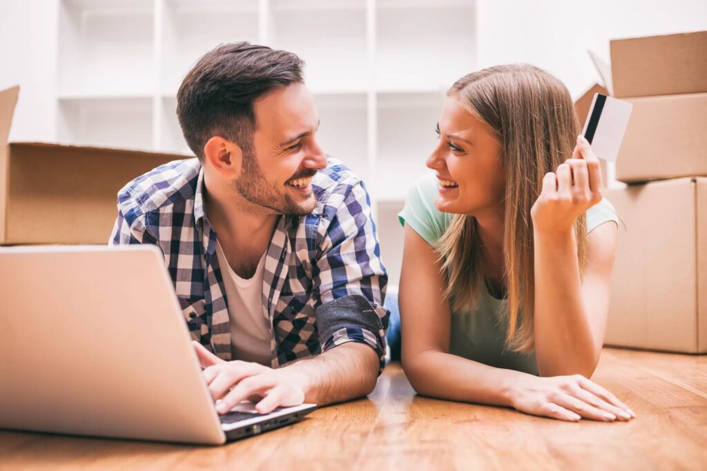 A couple smiling at each other while lying on the floor next to a laptop and boxe